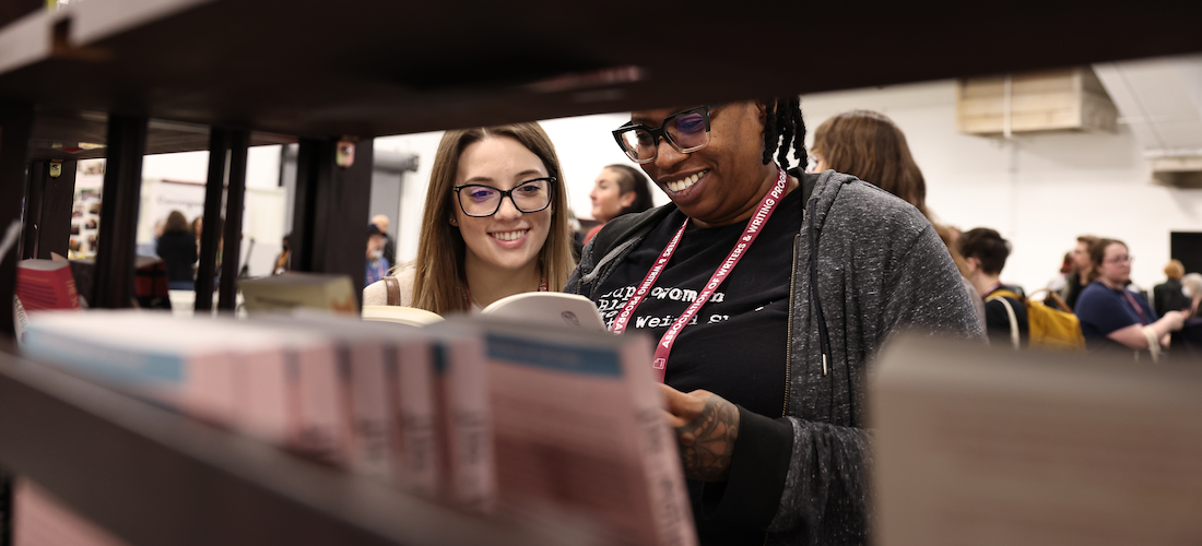 Two women reading looking at book, in front of a bookshelf.