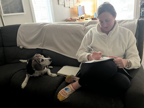 Person writing in notebook while sitting on couch next to a dog